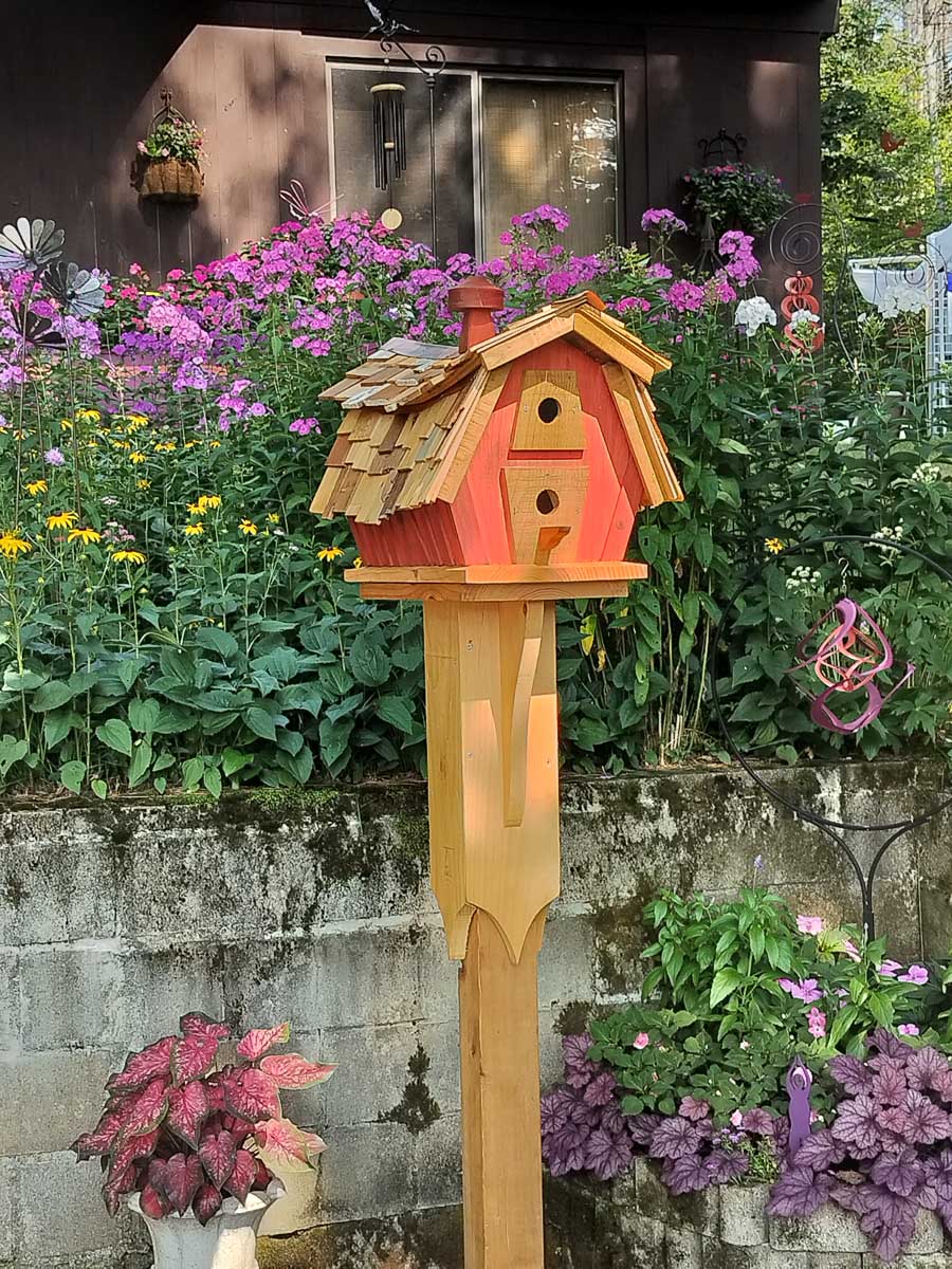 A Swayback style birdhouse stained orange standing in front of a purple and pink garden.